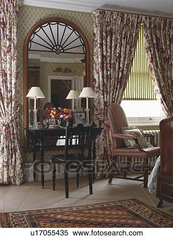 Stock Photography - Large mirror above antique table and chair in townhouse bedroom with patterned curtains and striped blind. Fotosearch