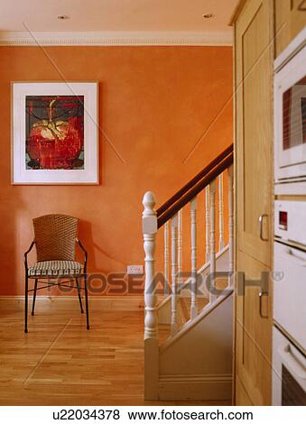 Stock Photo - Large picture above wicker and metal chair in orange hall with wooden flooring. Fotosearch