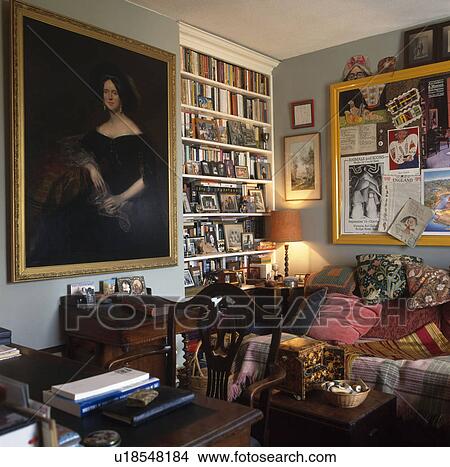 Picture - Large portrait painting in grey living room with bookshelves and lighted lamp beside sofa piled with cushions. Fotosearch