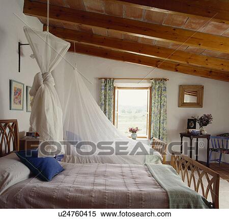 Stock Photography - Large white bedroom with a sloping wooden ceiling, and twin bed with mosquito nets . Fotosearch