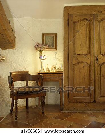 Stock Image - Lighted lamp on small table between antique wooden chair and old pine doors on built-in cupboard. Fotosearch