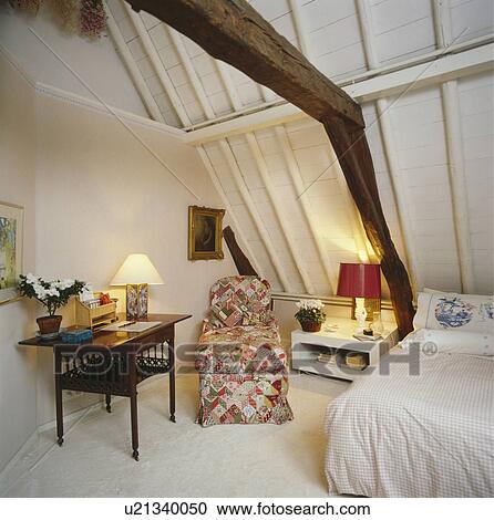 Stock Image - Lighted lamps and floral chaiselongue in white attic bedroom with sloping white wooden ceiling and white carpet. Fotosearch
