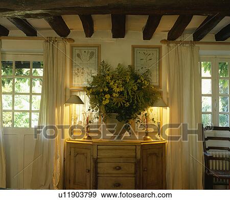 Lighted lamps and flower arrangement on cupboard between windows in white cottage living room View Large Photo Image Stock Photo - Lighted lamps and flower arrangement on cupboard between windows in white cottage living room. Fotosearch