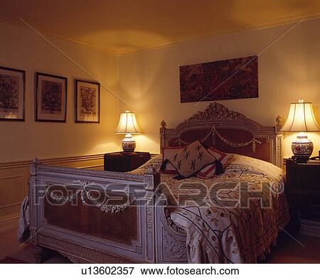 Stock Photo - Lighted lamps on either side of carved and painted antique bed in small traditional bedroom. Fotosearch