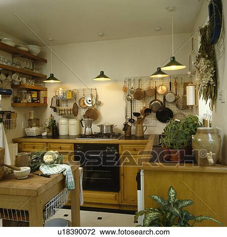 Stock Image - Lighted pendant lamps above oven and hob in wooden unit in small kitchen with pans on metal wall rack. Fotosearch