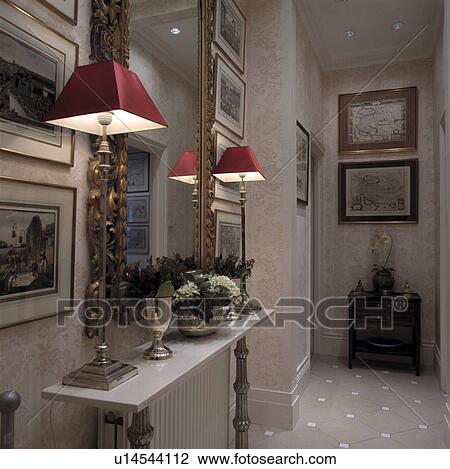 Stock Image - Lighted red lamps on shelf below gilt mirror in narrow beige hall with beige+white tiled floor. Fotosearch