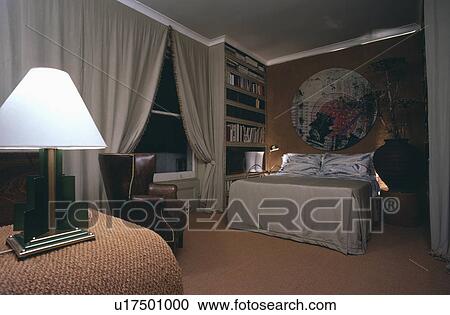 Stock Image - Lighted white lamp and grey curtains in bedroom with circular oriental picture above bed with grey bedcover. Fotosearch