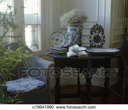 Stock Image - Lloyd Loom armchair beside antique table with blue+white china in traditional living room. Fotosearch