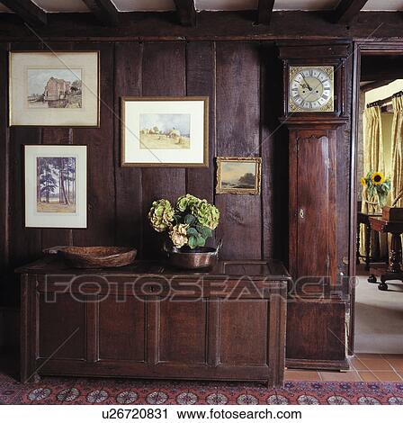 Stock Image - Longcase clock beside antique carved wooden chest in panelled country hall. Fotosearch