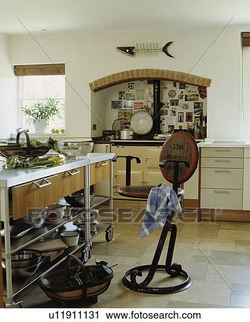 Stock Image - Metal and wood stool beside freestanding mobile metal island unit in modern white kitchen with cream Aga. Fotosearch