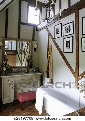 Stock Photo - Mirror above white dressing table in country bathroom with black+white photographs on beamed wall above white bath. Fotosearch