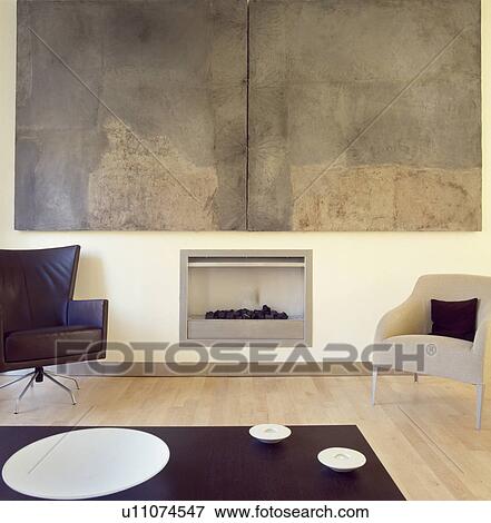Modern livingroom with cream armchair and black leather swivel armchair on either side of modern fireplace below large abstract painting by Hannah Biggs View Large Photo Image Stock Photo - Modern livingroom with cream armchair and black leather swivel armchair on either side of modern fireplace below large abstract painting by Hannah Biggs. Fotosearch