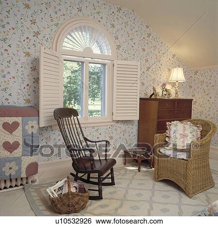 Stock Photograph - Oak rocking chair and wicker armchair in bedroom with floral wallpaper and white shutters on arched window . Fotosearch