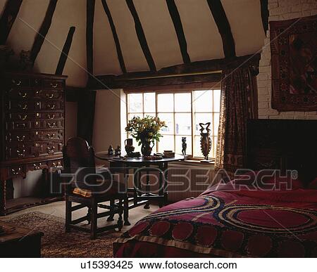 Oak table and chairs in window of country bedroom with patterned red bedcover and beamed apex ceiling View Large Photo Image Stock Photography - Oak table and chairs in window of country bedroom with patterned red bedcover and beamed apex ceiling. Fotosearch