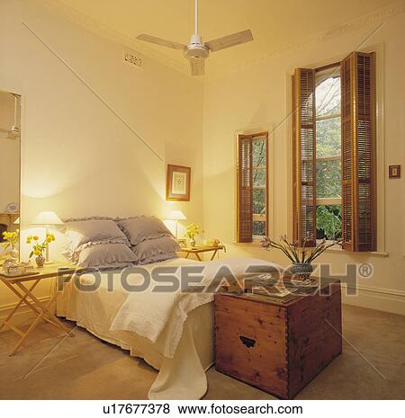 Stock Photo - Old pine chest below bed with blue pillows and cream quilt and linen in white bedroom with wooden plantation shutters. Fotosearch