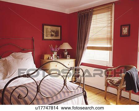 Picture - Ornate wrought iron bed in small red bedroom with beige curtain at window with slatted wooden blind. Fotosearch