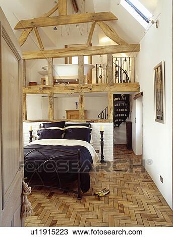 Stock Image - Parquet floor and black+white bedlinen on bed in openplan bedroom and mezzanine bathroom in barn conversion. Fotosearch