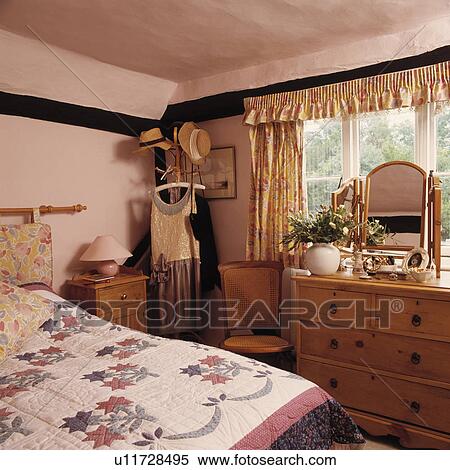 Stock Photography - Patchwork quilt of bed in pink cottage bedroom with pine chest-of-drawers in front of window with floral curtains. Fotosearch