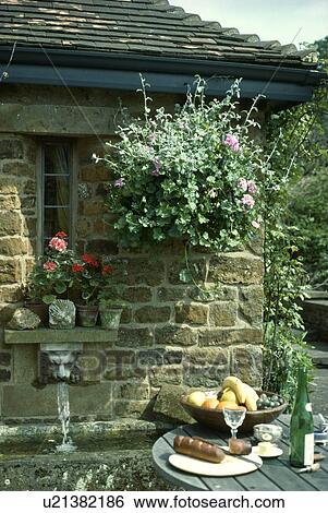 Stock Photograph - Patio table in front of cottage wall with hanging basket above lions head water fountain and stone sink. Fotosearch