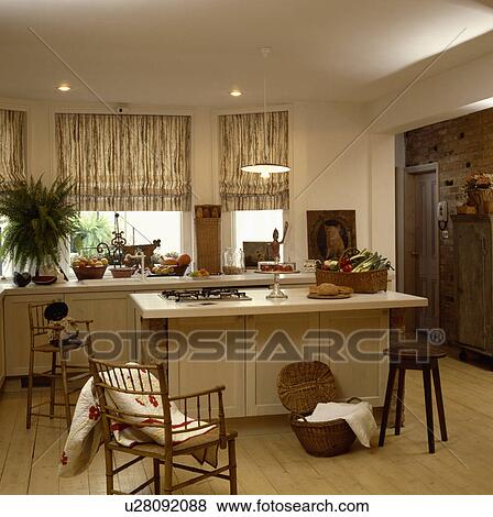 Patterned blinds in cream kitchen with antique chairs and wooden stool beside rectangular island unit View Large Photo Image Stock Photo - Patterned blinds in cream kitchen with antique chairs and wooden stool beside rectangular island unit . Fotosearch