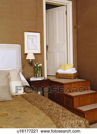 Stock Image - Patterned gold linen on bed in light brown bedroom with storage drawers in steps leading to white door. Fotosearch
