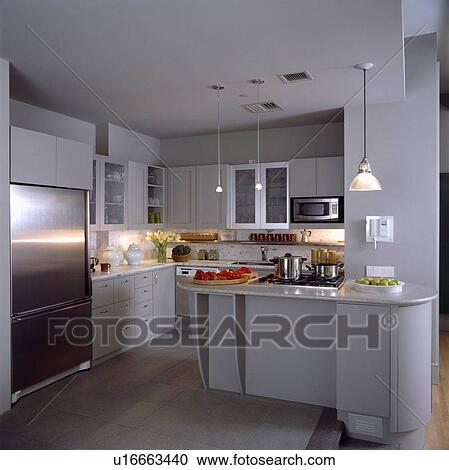 Stock Image - Pendant lighting in modern white kitchen with hob built into worktop large stainless steel freestanding fridge . Fotosearch