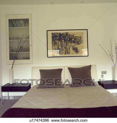 Stock Photograph - Picture above bed with brown velvet cushions and neutral bedlinen in modern bedroom with frosted glass door on flush-fitted bookshelves. Fotosearch