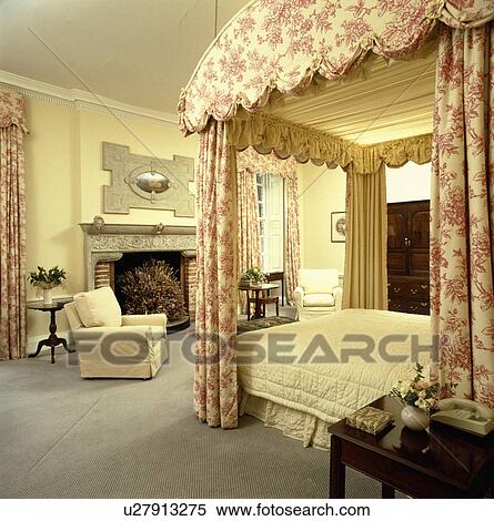 Stock Photography - Pink Toile de Jouy drapes and cream quilt on fourposter bed in large pastel yellow country bedroom with grey carpet. Fotosearch