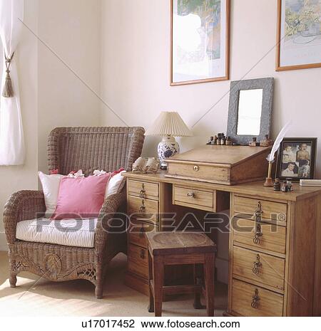 Stock Image - Pink & white cushions on wicker armchair beside old pine desk and stool in small study. Fotosearch