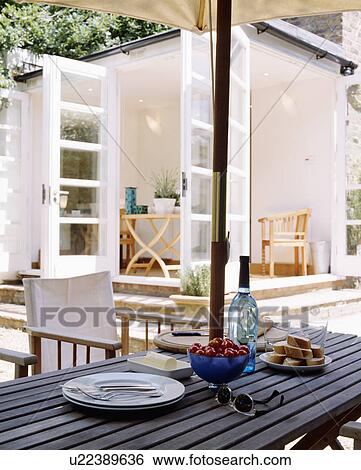 Plates and food on wooden patio table with white director's chairs and umbrella and view of modern white livingroom extension View Large Photo Image Stock Photograph - Plates and food on wooden patio table with white director's chairs and umbrella and view of modern white livingroom extension. Fotosearch