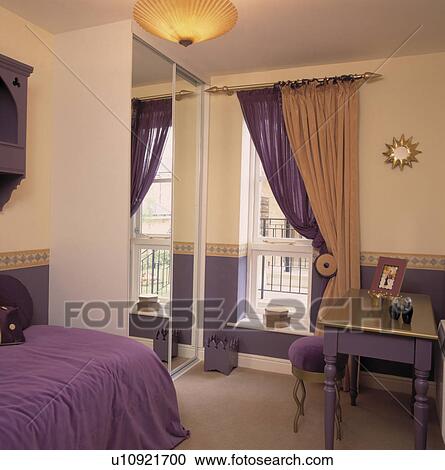 Stock Image - Purple and neutral curtains at window in bedroom with fitted mirrored wardrobe and purple bedling and table with gilt top. Fotosearch