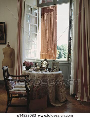 Stock Image - Quilted cloth on circular table in front of open window with shutters and striped curtains in French country bedroom. Fotosearch