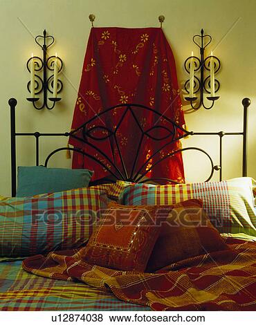 Red and gold wall-hanging behind black iron bed piled with colourful checked pillows and red throw and cushions View Large Photo Image Stock Photo - Red and gold wall-hanging behind black iron bed piled with colourful checked pillows and red throw and cushions. Fotosearch