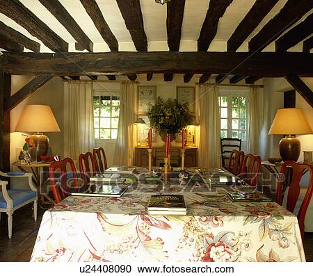 Stock Image - Red candles on floral tablecloth in country living room with beamed ceiling and lighted lamps. Fotosearch