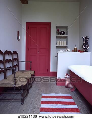 Stock Photograph - Red door in white bathroom with antique rush-seated settle and red+white striped rug beside clawfoot bath. Fotosearch