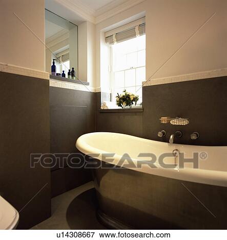 Stock Photo - Roll-top high-sided bath with central taps in modern white bathroom with dark grey wall tiles. Fotosearch