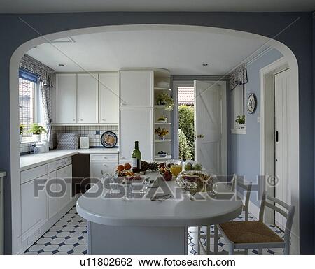 Stock Image - Rush-seated stools at island breakfast bar in pastel blue kitchen with white fitted cupboards. Fotosearch