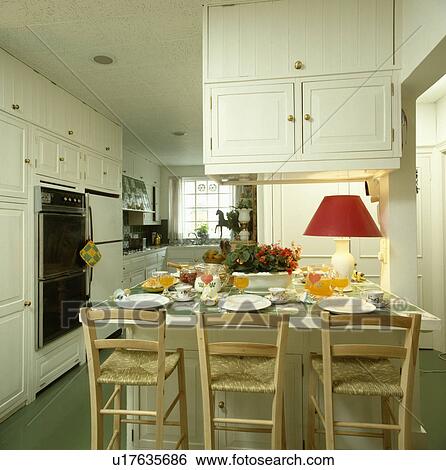 Rush-seated wooden chairs and red lamp on table with place settings in white kitchen View Large Photo Image Stock Photograph - Rush-seated wooden chairs and red lamp on table with place settings in white kitchen. Fotosearch