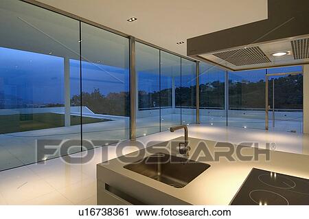 Stock Image - Sink in island unit in modern Spanish kitchen with glass walls and view of terrace at night. Fotosearch