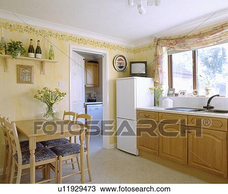 Stock Image - Small television on top of fridge freezer in pastel yellow kitchen dining room with fitted units and wooden table. Fotosearch