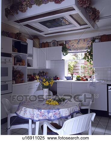 Stock Photography - Stained glass skylight above patterned cloth on circular table with white chairs in traditional white kitchen. Fotosearch