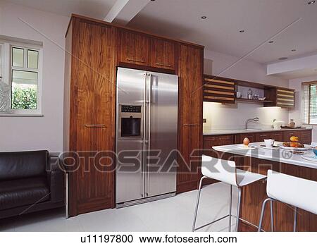 Stock Image - Stainless steel American-style refrigerator in modern kitchen with white stools at breakfast bar and fitted walnut cupboards. Fotosearch