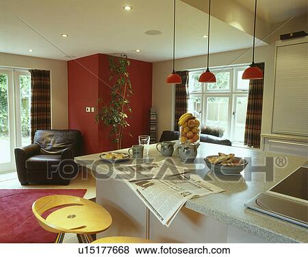 Stock Photo - Stools with newspaper and breakfast on bar on island unit in modern white kitchen with leather armchair against red wall. Fotosearch