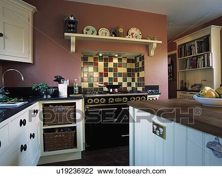 Stock Image - Storage baskets in fitted unit beside range oven below shelf and multicoloured tiles in dark pink and white kitchen. Fotosearch