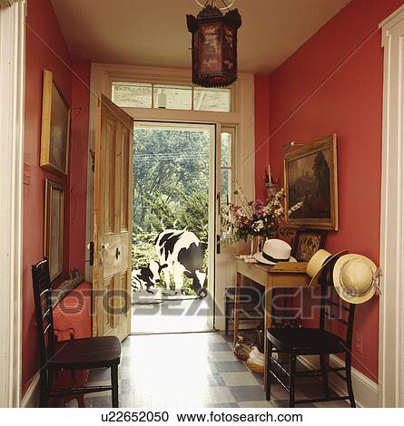 Stock Image - Straw hats on antique chairs in red hall with blue+white checked floor and open door with view of model cows . Fotosearch
