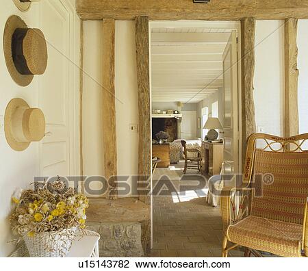 Stock Image - Straw hats on wall in beamed cottage hall with wicker chair beside doorway with view of living room. Fotosearch