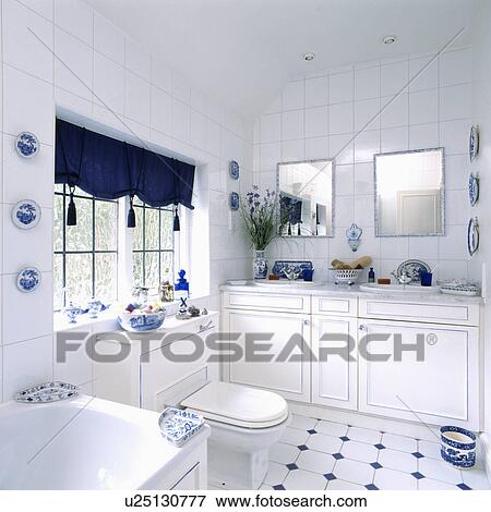 Stock Photo - Tasselled blue blind in white bathroom with mirrors above double basins and white and blue tiled floor. Fotosearch