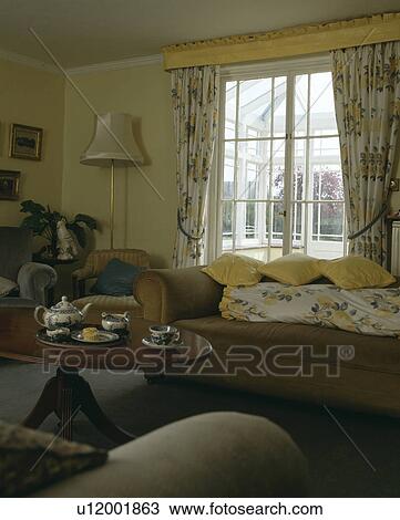 Stock Image - Teapot and cups on small circular table in front of sofa in livingroom with yellow floral curtains. Fotosearch