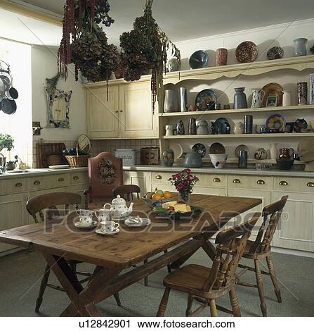 Stock Image - Teaset on wooden table in neutral kitchen with jugs and bowls on shelves above worktop. Fotosearch
