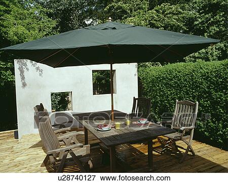 Stock Photo - Umbrella and wooden furniture on decked patio in front of white wall. Fotosearch